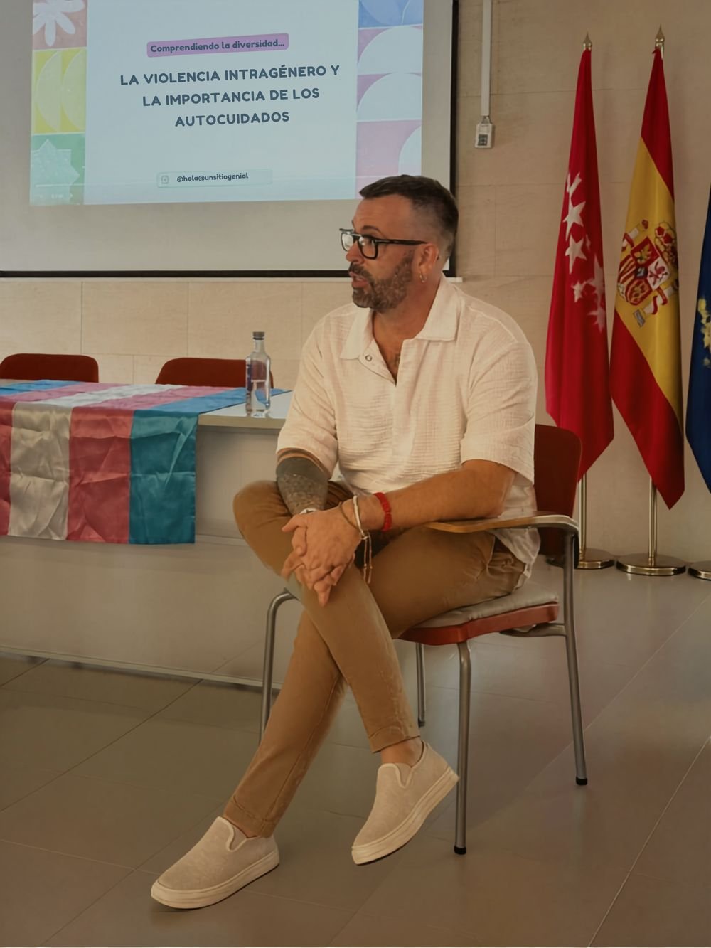 Fran Pardo sentado durante una charla sobre violencia intragénero, con una bandera trans sobre la mesa y la presentación proyectada detrás
