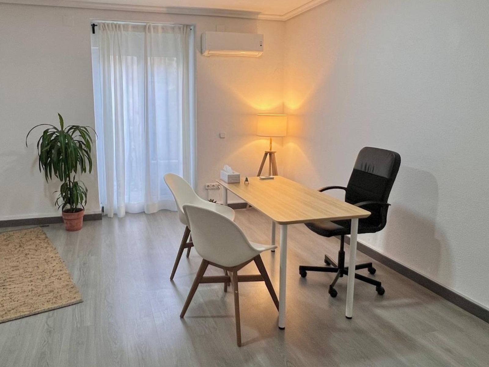 Sala del despacho con mesa de trabajo, dos sillas para quien viene, ventana con visillos, lámpara encendida y una planta